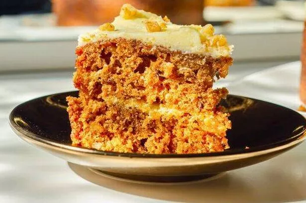 Close-up Image of a slice of Carrot Cake, White White Creamy Frosting on Top and Sprinkled With Nuts, Resting on A Black Saucer.
