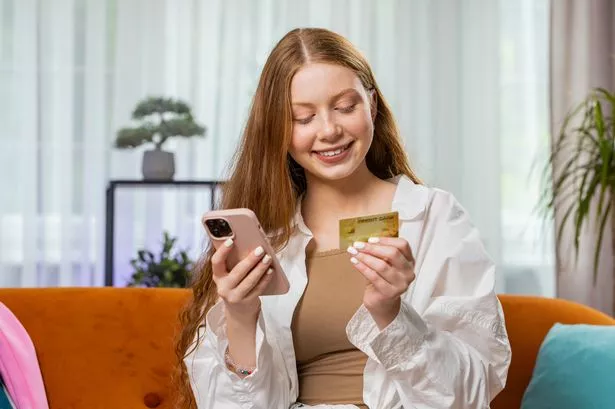 Teenager Girl USING Credit Bank Card and Smartphone while Transferring Money, Online Cashless Shopping, ORDER FOOD DELIVERY AT HOME INDOORS. HAPPY CHILD SITTING IN ROOM ON COUCH