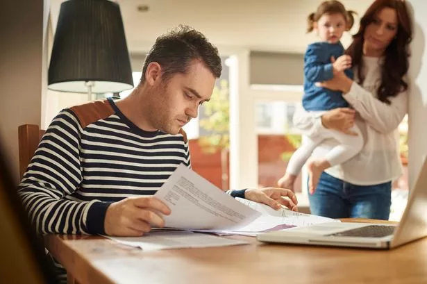 A Young Husband and Father Sites at A Dining Table Looking Through the Household Bills and Looks World. His Wife and Young Daughter Stand in the background looking on sympathetically.