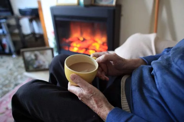 Pensioner holding cup of coffee sitting in front of an open fire.