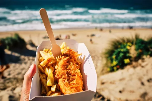 POV hand holding a box of Fish and Chips ( Cod and Chips ) above Fistral Beach, Newquay, Cornwall on a sunny September day.