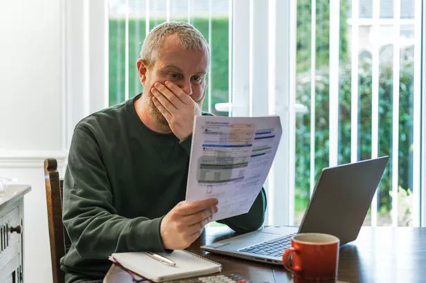 Portrait of a mid adult man in his 30s checking his energy bills at home. He has a worried expression and touches his face with his hand while looking at the bills. He is surrounded by documents, his laptop and a calculator.