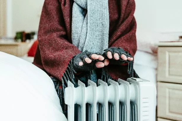A retired senior man in his 70s sits at home inside his cold house in winter. It is so cold that he is wrapped up in warm winter clothing, including gloves and a blanket, and is holding his hands over an electric heater for some extra warmth and comfort.